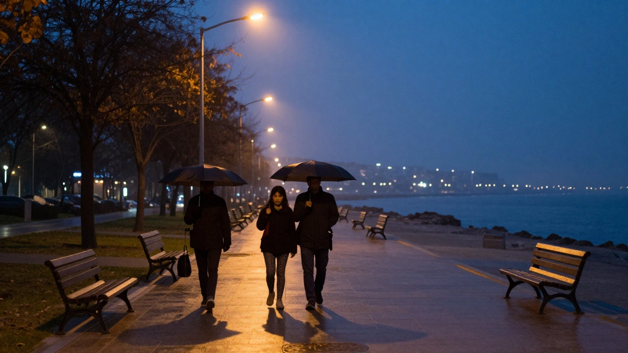Kartal Sahili'nde gölgeler uzarken, iki kişi yağmurdan korunmak için şemsiyelerle yürüyor.